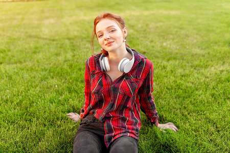 一个漂亮的红头发的学生与雀斑穿着一个红色的格子衬衫, 耳机坐在 cheba 之间的草坪上。学生休闲