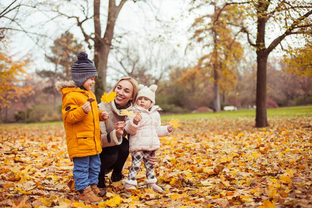 一个年轻漂亮的母亲和两个小孩在秋季公园散步。妈妈和两个小孩玩耍。温暖的冬天。明亮的秋天