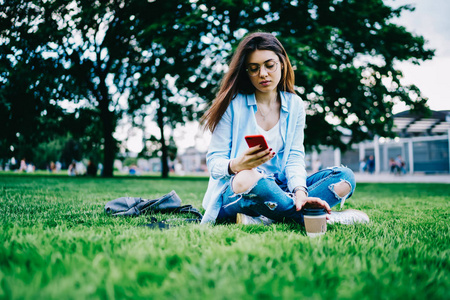 眼镜上的年轻沉思的妇女阅读收入信息智能手机坐在公园草在自由时间, 时髦女孩在社交网络中使用蜂窝和4g 连接的更新配置文件户外活动