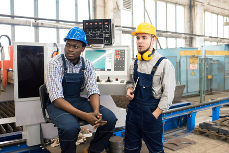 英俊的胡子机操作员和他的非洲裔美国人穿着 hardhats 和工作服站在 Cnc生产部内部的背景上讨论工作过程