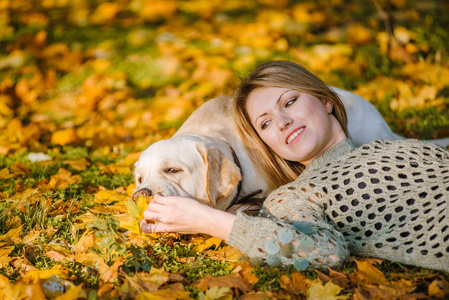 一个美丽的金发女郎躺在公园里的黄树叶上, 和她的拉布拉多犬玩耍。