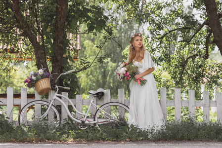 美丽的年轻新娘与婚礼花束和自行车站在栅栏附近, 看着远离