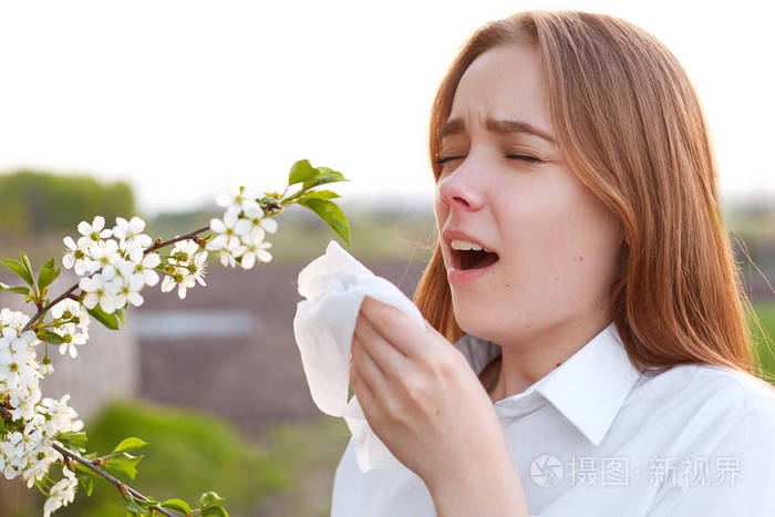 漂亮的年轻女子擤鼻涕 打喷嚏 站在盛开的树前 对开花过敏 拿手帕