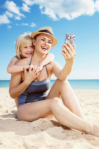 微笑的年轻母亲和孩子泳装拍照与智能手机在海滩上