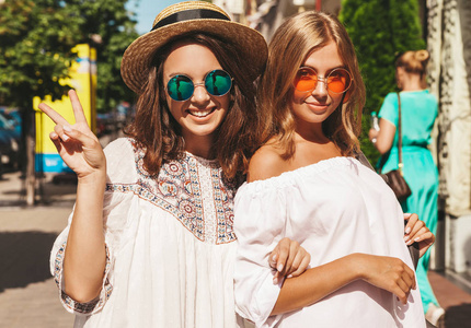 时尚肖像的两个年轻时尚的嬉皮黑发和金发女郎在夏季晴天穿着白色时髦的衣服摆在街头背景。不化妆