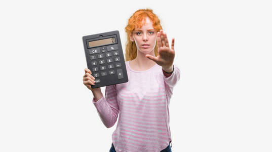手持大计算器的年轻红头发的女人用张开的手做停止标志以严肃和自信的表达, 防御手势