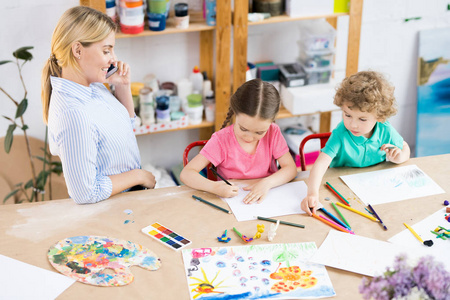 孩子们坐在木桌上, 用五颜六色的铅笔画一幅画, 老师在手机上聊天, 看着她的瞳孔如何工作。