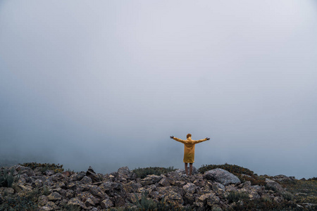 女性穿着黄色雨衣, 牛仔裤短裤站在山顶上, 地平线上有山峰的景色。景观。自然