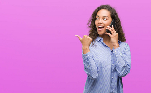 年轻的西班牙裔妇女使用智能手机指向和显示与拇指到一边, 快乐的脸微笑