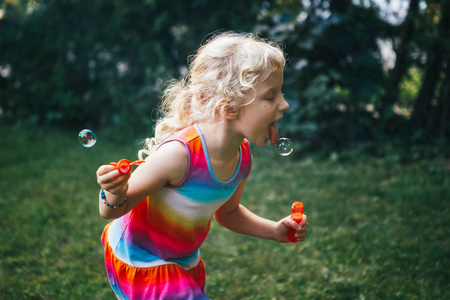 孩子试图抓住吃肥皂气泡与舌头。在夏天日落时在家后院玩的可爱滑稽的金发白种女孩的坦率的肖像。真正真实的快乐童年时刻