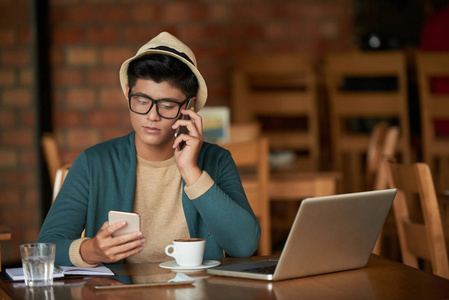 年轻的亚裔男子打电话和手持手机在桌子上与笔记本电脑