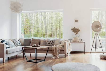 在温暖的起居室内用白色的墙壁, 用一个大玻璃窗在米色的角落沙发上打印图案枕头