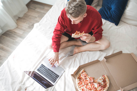 穿着红色运动衫的年轻人坐在一张床上, 上面放着一个披萨盒和一台笔记本电脑。学生坐在床上的 dmao 吃比萨饼和使用笔记本电脑。带