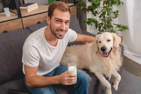 高视角的微笑的年轻人与玻璃牛奶和可爱的金色猎犬狗看着相机