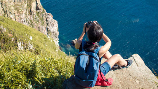 年轻女子摄影师坐在悬崖边上, 拍一张风景照片。美丽的岩石海岸