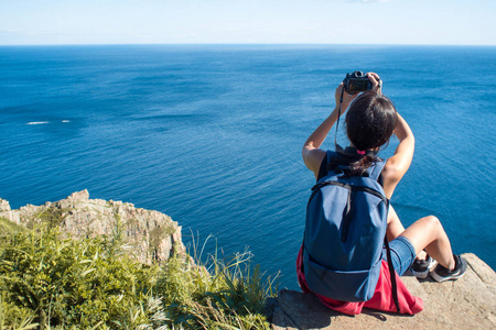 年轻女子摄影师坐在悬崖边上, 拍一张风景照片。美丽的岩石海岸