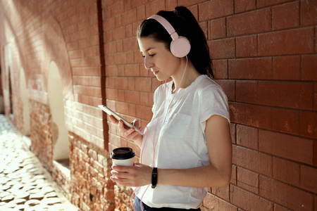 年轻漂亮的白种女人, 手里拿着智能手机和咖啡杯, 独自站在砖墙旁边。清晨跑步, 醒来, 听音乐, 粉红色的耳机, 检查电子邮件