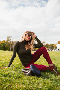 快乐时尚的女人戴着太阳镜坐在绿色的草地上摆在公园里