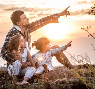 幸福美满的家庭, 爸爸和两个小可爱的女儿在日落时坐在一座高高的美丽的山上, 家庭关系和友谊的概念