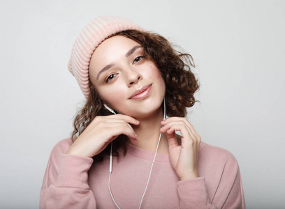 一个美丽的女孩在耳机的肖像听音乐和微笑, 而站在白色的背景, 粉红色的运动衫和帽子。生活方式概念
