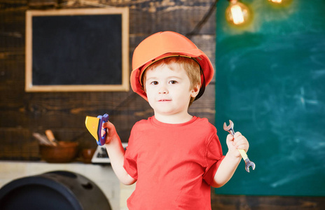 幼儿在保护硬帽子, 头盔在家里的车间。童年的概念。小男孩扮演杂工。儿童可爱玩扳手和玩具锯, 作为建设者或修理工, 修理或就要手工