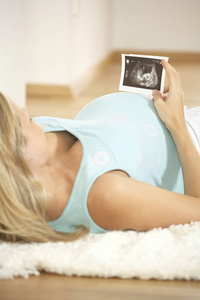 在家里躺在地板上的年轻孕妇在腹部持有超声波图像