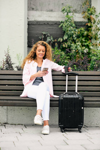 在车站的长凳上坐着的女人的肖像起诉智能手机。她的行李旁边