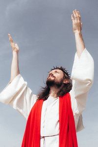 耶稣在长袍红窗扇和荆棘王冠上的低角观, 站立着举起手对抗灰色的天空