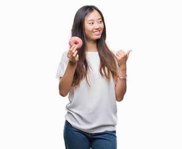 年轻的亚洲妇女吃甜甜圈在孤立的背景下指向和显示与拇指到一边与快乐的脸微笑