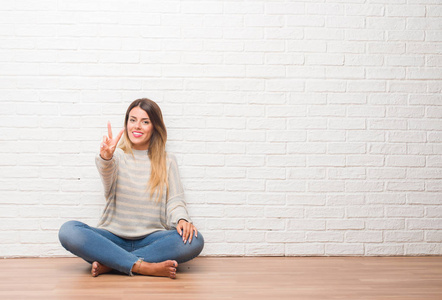年轻的成年女子坐在地板上的白色砖墙在家里微笑着看着相机显示手指做胜利的标志。第二个