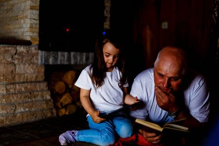 祖父和孙女读书和使用平板在壁炉附近
