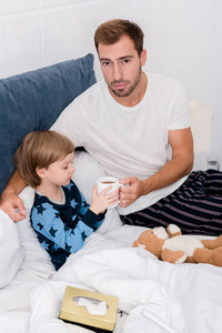 年轻的父亲给他的儿子在床上喝一杯热饮料, 看着相机