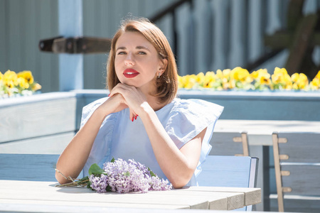 穿着淡蓝色连衣裙的年轻迷人女子的户外肖像坐在夏日咖啡馆的桌旁, 带着新鲜的丁香花