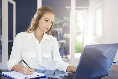 认为年轻的女商人坐在笔记本电脑前做笔记, 在商业项目上工作