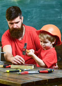 父亲用胡子教小儿子用工具, 锤击, 黑板上的背景。男孩, 孩子忙着保护头盔学习用锤子和爸爸在一起。小助手概念