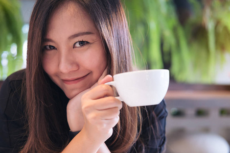 在现代咖啡馆和绿色自然背景下, 一个美丽的亚洲女人手持一只白色的杯子, 喝着热咖啡, 感受着快乐的特写图像
