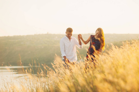 微笑欢快的年轻可爱的夫妇走在湖附近的山丘上日落和牵手