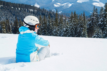 穿着蓝色西装头盔和眼镜的妇女坐在山顶上滑雪板附近的雪地上。