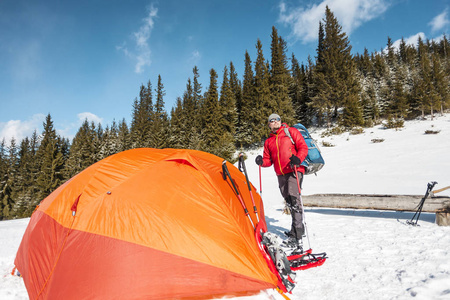 男子在帐篷附近的冬天。游客在极端的条件。雪地鞋, 徒步竿和背包。在冬山露营。橙色帐篷在雪里