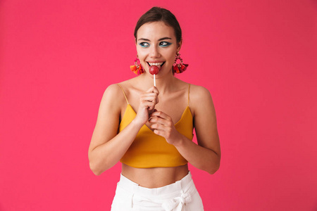 欧洲内容女性肖像20s 戴着耳环微笑着吃着粉红色背景下的棒糖