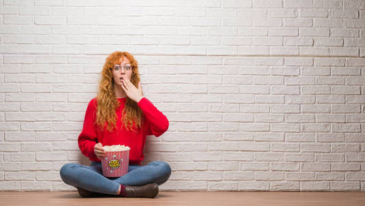 年轻的红头发女人坐在砖墙上吃爆米花捂着嘴用手震惊的错误, 表情的恐惧, 在沉默中害怕, 秘密概念