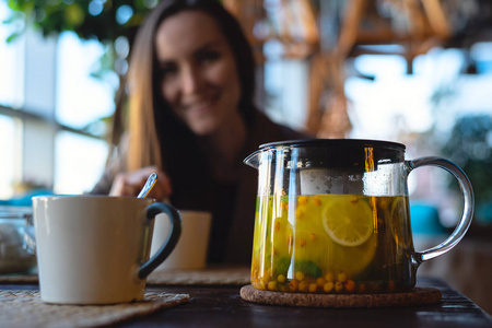 在一个微笑的女孩的背景下, 一个杯子和一个带有柠檬和香草的沙棘茶的玻璃茶壶特写