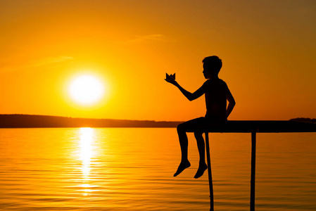一个小男孩坐在桥上, 晚上在日落时手里拿着纸质的折纸船。一个男孩在河边玩他的玩具。