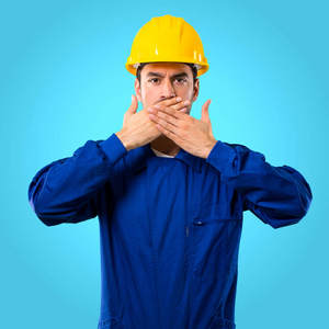年轻的工人用头盔捂住嘴, 用手说一些不合适的东西。在蓝色背景下不能说话