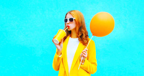 时尚肖像女人喝果汁在彩色蓝色背景上持有橙色热气球