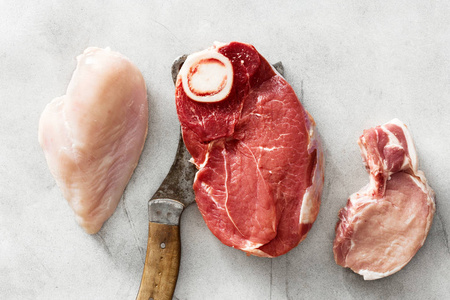 各种生肉, 牛肉肉骨, 猪肉和鸡柳在灰色的石头背景与屠夫刀, 顶部视图