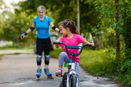 年轻的母亲轮滑与小女儿骑自行车在公园