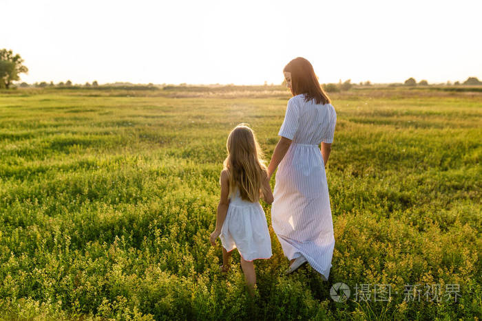 母亲和女儿牵手走在绿色的草地上, 日落的天空背景下