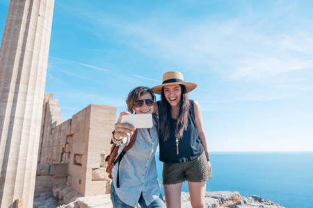 两个年轻美丽快乐的女人女性旅行女友在希腊的海洋和古遗址的背景下做自拍