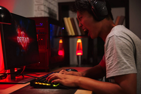 亚洲时态愤怒的游戏玩家男孩在玩电子游戏时丢失的肖像戴着耳机和使用背光彩色键盘的电脑在黑暗的房间里